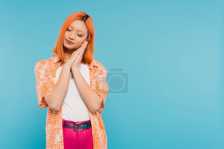 sleepy asian woman, young model with dyed red hair holding hands near face and posing with closed eyes on vibrant blue background, generation z, casual attire, tired, fatigue, exhausted 
