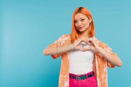 love sign, happy face, cheerful young asian woman with dyed red hair smiling and showing heart with hands on vibrant blue background, generation z, casual attire, looking at camera, young culture 
