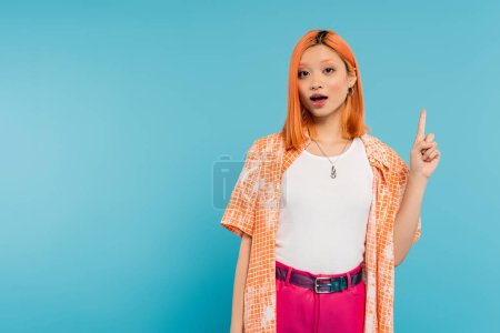 pointing with finger, amazed face, young asian woman with dyed hair looking at camera and showing something on blue background, generation z, casual attire, young culture 