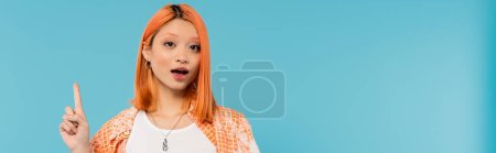 wskazując palcem, zdumiona twarz, młoda Azjatka z farbowanymi włosami pokazująca coś na kamerze na niebieskim tle, pokolenie z, luźny ubiór, młoda kultura, ekspresyjny, baner internetowy 