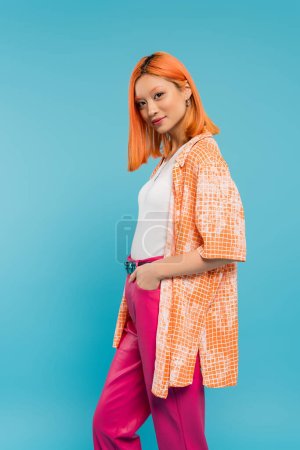 Hand in Tasche, hübsche junge Asiatin mit gefärbten Haaren, die in orangefarbenem Hemd vor blauem Hintergrund posiert, in die Kamera blickt, rosa Hose, Generation Z, moderner Stil 
