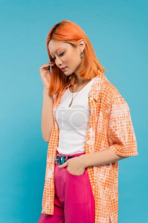 Hand in Tasche, sinnliche junge Asiatin mit gefärbten Haaren, die im orangefarbenen Hemd vor blauem Hintergrund posiert, wegschaut, rosa Hose, Generation Z, moderner Stil 