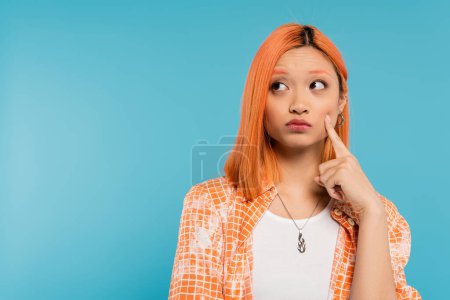 asiatische Frau denkt und sieht weg, junges Modelmodel berührt Wange mit Finger auf blauem Hintergrund, nachdenklich, orangefarbenes Hemd, Generation z, leuchtende Farben, zweifelhaftes Gesicht 