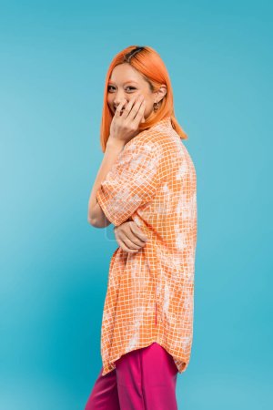positivity, shy and young asian woman with dyed hair standing in orange shirt and covering mouth with hand while smiling on blue background, looking at camera, joyful, happiness, generation z 