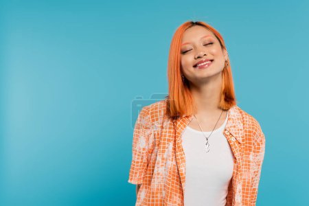 joyful face, radiant smile, young asian woman with dyed hair standing with closed eyes in orange shirt and smiling on blue background, casual attire, happiness, freedom, cheerful attitude 