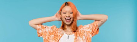 positividad y felicidad, joven mujer asiática con el pelo teñido de pie con los ojos cerrados en camisa naranja y sonriendo sobre fondo azul, atuendo casual, libertad, actitud alegre, tatuaje, bandera 