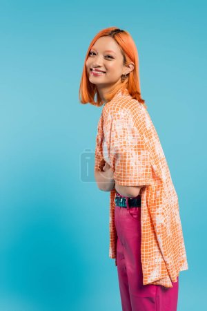 sonrisa radiante, joven mujer asiática con el pelo teñido de pie con los brazos cruzados, en camisa naranja y sonriendo sobre fondo azul, atuendo casual, libertad, actitud alegre, mirando a la cámara, positividad 