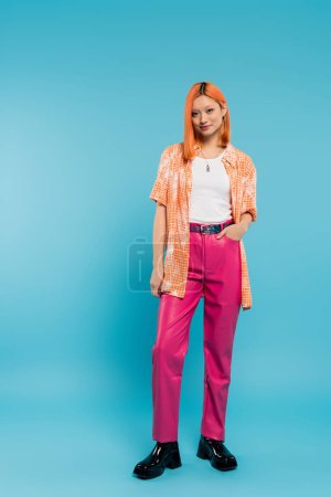 main dans la poche, souriante et jeune femme asiatique avec les cheveux teints debout en chemise orange et posant sur fond bleu, regardant appareil photo, pantalon rose, génération z, style moderne, pleine longueur 