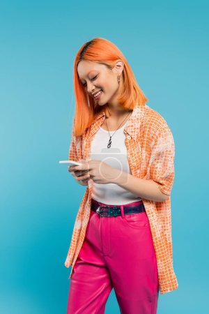 réseautage social, femme asiatique joyeuse avec des messages capillaires teints, en utilisant un smartphone, debout sur fond bleu, sourire, chemise orange, tenue décontractée, natif numérique, génération z 