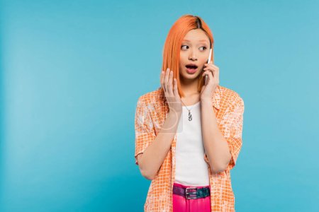 surprised asian woman during phone call, young model with dyed hair standing with opened mouth and talking on smartphone on blue background, looking away, emotional, shocked face 
