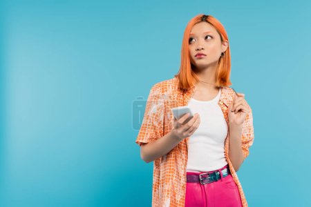 social media influencer, pensive asian woman with dyed hair holding smartphone and looking away on blue background, mobile phone, youth culture, digital age, generation z