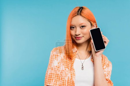 social media influencer, happy and young asian woman with dyed hair holding smartphone with blank screen on blue background, mobile phone, youth culture, digital age, generation z 