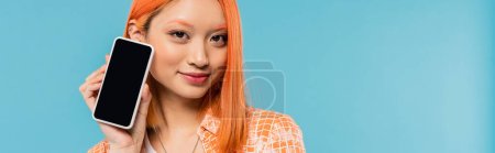 social media influencer, joyful and young asian woman with dyed hair holding smartphone with blank screen on blue background, mobile phone, youth culture, digital age, generation z, banner 