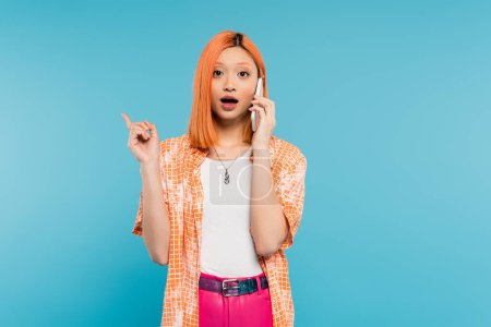 Photo for Phone call, advertisement, young asian woman with dyed hair pointing with finger and talking on smartphone on blue background, mobile phone, youth culture, digital age, generation z, surprised face - Royalty Free Image