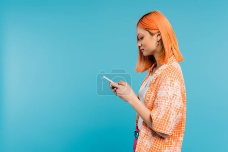 Photo for Social media influencer, young asian woman with dyed hair using smartphone on blue background, mobile phone, youth culture, digital age, generation z, messaging, side view - Royalty Free Image