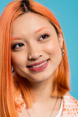 portrait, joyeuse femme asiatique aux cheveux courts et teints, maquillage naturel et boucles d'oreilles cerceau regardant loin sur fond bleu, chemise orange, génération z, mode, visage heureux, sourire radieux 