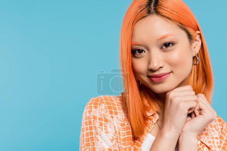 retrato, actitud alegre, joven mujer asiática con pelo corto y teñido, maquillaje natural y pendientes de aro mirando a la cámara sobre fondo azul, camisa naranja, generación z, cara feliz, sonrisa radiante 