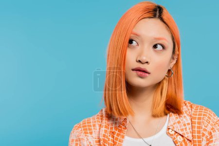 retrato, pensando y mirando hacia otro lado, pensativo joven mujer asiática con pelo corto y teñido, maquillaje natural y pendientes de aro sobre fondo azul, camisa naranja, generación z, labio mordedor 