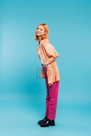 posing with hand in pocket, joyful and young asian woman with dyed hair standing in casual wear on blue background, looking at camera, pink pants, generation z, modern style, full length 