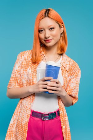 boire dans les mains, heureux asiatique et jeune femme aux cheveux roux tenant tasse en papier et en regardant la caméra sur fond bleu, tenue décontractée, génération z, culture du café, boisson chaude, tasse à usage unique 