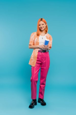 Photo for Cheerful asian and young woman with red hair holding paper cup and looking at camera on blue background, casual attire, generation z, coffee culture, hot drink, single use cup, full length - Royalty Free Image