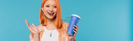 positivité, café à emporter, heureuse asiatique et jeune femme aux cheveux roux tenant une tasse en papier et regardant la caméra sur fond bleu, tenue décontractée, génération z, culture du café, boisson chaude, étonné, bannière