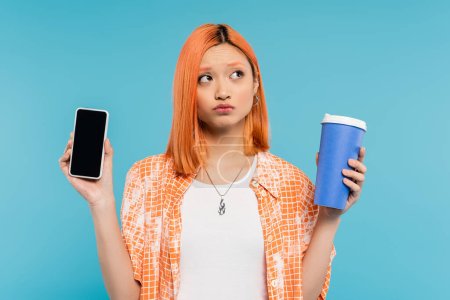 smartphone et café, asiatique et jeune femme aux cheveux roux tenant tasse en papier et téléphone mobile sur fond bleu, tenue décontractée, culture du café, génération z, lèvres boudantes, penser, écran vierge