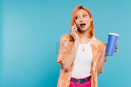phone call, emotional asian and young woman with red hair holding takeaway drink and talking on smartphone on blue background, casual attire, generation z, coffee culture, amazed