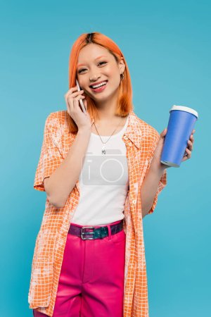 llamada telefónica, mujer asiática alegre con pelo rojo sosteniendo café para ir en taza de papel y hablando en el teléfono inteligente sobre fondo azul, atuendo casual, generación z, cultura del café, bebida caliente 