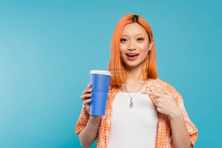 Positivität, glückliche asiatische und junge Frau mit roten Haaren, die auf Pappbecher zeigt und in die Kamera auf blauem Hintergrund schaut, lässige Kleidung, Generation z, Kaffeekultur, Heißgetränk, erstaunt