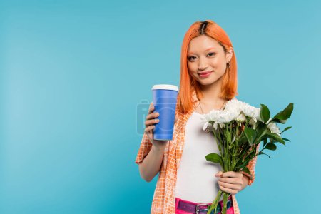 positividad, feliz asiática y joven mujer con el pelo rojo sosteniendo café para ir en taza de papel y ramo de flores sobre fondo azul, atuendo casual, generación z, cultura del café, bebida caliente 