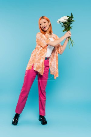 Positivität, erstaunte asiatische Frau mit roten Haaren, die weiße Blumen auf blauem Hintergrund hält, lässige Kleidung, Generation z, Blumenstrauß, Frühlingsstimmung, glückliches Gesicht, Gen z, Jugendkultur, volle Länge
