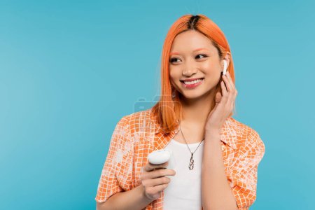 summer happiness, music lover, young and trendy asian woman with dyed hair, in orange shirt holding case, adjusting wireless earphone and looking away on blue background, generation z