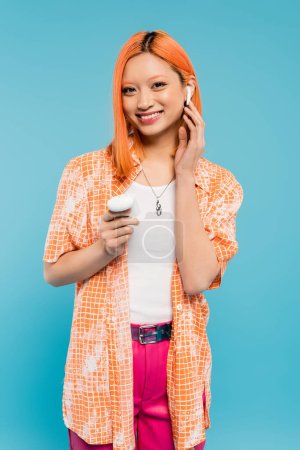 fröhliche Musikliebhaberin, junge Asiatin in orangefarbenem Hemd, mit gefärbten roten Haaren, die den drahtlosen Kopfhörer einstellen und in die Kamera schauen, während sie die Tasche auf blauem Hintergrund hält, Generation z
