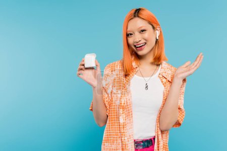 amazed asian woman with happy face and colored red hair wearing stylish orange shirt and listening music in wireless earphone while standing with case on blue background, youth culture