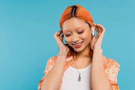 young and happy asian woman with colored red hair listening music in wireless earphone, fixing red colored hair and smiling on blue background, generation z, summer vibes