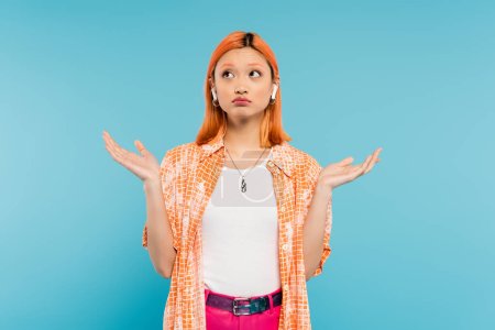 Téléchargez les photos : Jeune femme asiatique découragée en chemise orange, avec des cheveux roux teints écoutant de la musique dans un écouteur sans fil, montrant un geste haussant les épaules et regardant vers le haut sur fond bleu, génération z - en image libre de droit