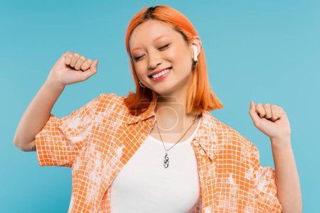 Glück, Musikliebhaberin, junge Asiatin mit gefärbten roten Haaren, im trendigen orangefarbenen Hemd und drahtlosem Kopfhörer gestikulierend mit geschlossenen Augen auf blauem Hintergrund, Generation-Z-Lifestyle, Sommerstimmung