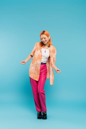 Sommerfreizeit, Musikliebhaberin, überglückliche Asiatin in bunten Kleidern und kabellosen Kopfhörern auf blauem Hintergrund, gefärbte Haare, orangefarbenes Hemd, rosa Hose, Gen z Mode