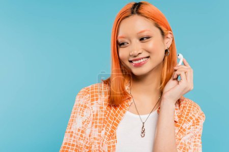 young and happy asian fashion model with colored red hair, in stylish orange shirt holding wireless earphone while standing on blue background, youth culture, generation z