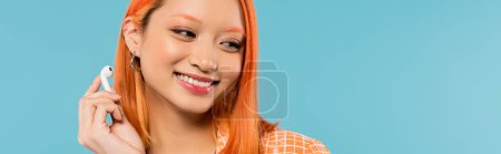 Glück und Freizeit, Porträt einer entzückenden asiatischen Frau mit strahlendem Lächeln und gefärbten roten Haaren mit drahtlosem Kopfhörer auf blauem Hintergrund, sommerliche Stimmung, Generation z, Banner