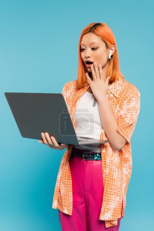 staunende asiatische Frau mit rot gefärbten roten Haaren, die die Hand in der Nähe des offenen Mundes hält und auf den Laptop auf blauem Hintergrund schaut, jugendliche Mode, orangefarbenes Hemd, freiberuflicher Lebensstil 