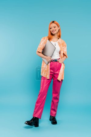 Foto de Felicidad y optimismo, longitud completa de mujer asiática pelirroja en camisa naranja sosteniendo portátil y de pie con la mano en el bolsillo de los pantalones de color rosa sobre fondo azul, joven freelancer, generación z - Imagen libre de derechos