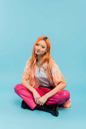 happy face, positive emotion, redhead and stylish asian woman sitting with crossed legs and looking at camera on blue background, orange shirt, pink pants, summer vibes