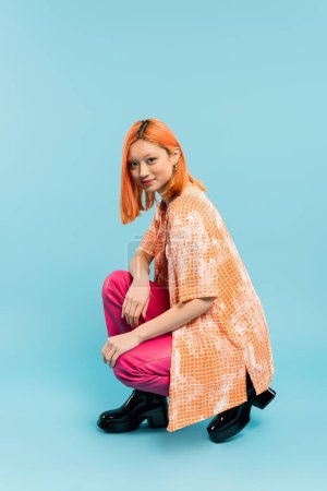 positive emotion, young asian woman with radiant smile looking at camera while posing on haunches on blue background, casual attire, orange shirt, pink pants, full length, generation z