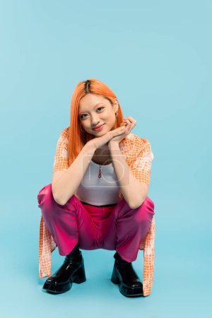 young and stylish asian woman with pleased face expression posing on haunches and looking at camera on blue background, colored red hair, casual attire, orange shirt, pink pants, full length