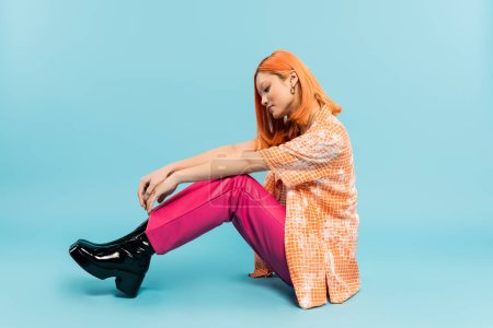 Photo for Side view of young and stylish asian woman in orange shirt and pink pants sitting on blue background, dyed red hair, summer fashion, youthful style, full length, generation z - Royalty Free Image