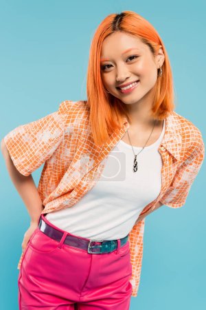 Foto de Mujer asiática alegre y de moda con el pelo rojo de color y sonrisa radiante posando en camisa naranja y mirando a la cámara en el fondo azul, feliz verano, generación z estilo de vida - Imagen libre de derechos