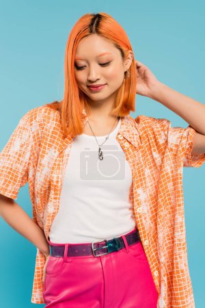 Foto de Mujer asiática joven y complacida con el pelo rojo de color rojo posando con la mano en la cadera sobre fondo azul, camisa naranja, pantalones rosados, moda juvenil, estilo veraniego, generación z - Imagen libre de derechos