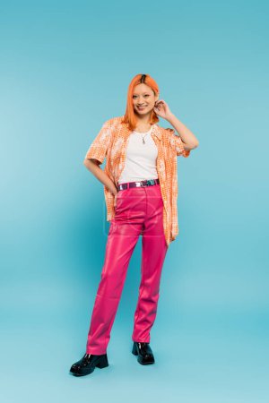 full length of young and redhead asian woman in trendy casual attire standing with hand on hip and smiling at camera on blue background, positive emotion, orange shirt, pink pants, youth culture